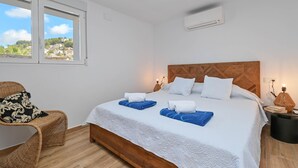 Ein Schlafzimmer mit einem hölzernen Kopfteil, einem großen Bett mit weißer und blauer Bettwäsche, einem Korbstuhl und Blick auf eine Stadt durch das Fenster.
