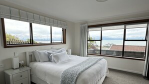 A bedroom with a large bed, a nightstand, a window with a view of the sea, and a ceiling fan.