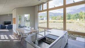 A modern kitchen with a large island, a dining area with a white table and chairs, and large windows offering a view of mountains and greenery. - Echo Rocks (Hakuba)