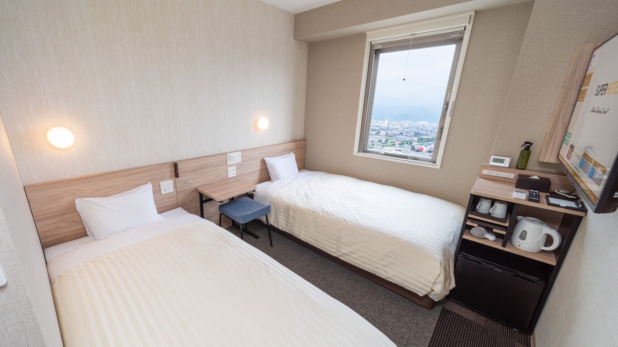 A hotel room with two beds, a desk, a window with a city view, a minibar, and a television.