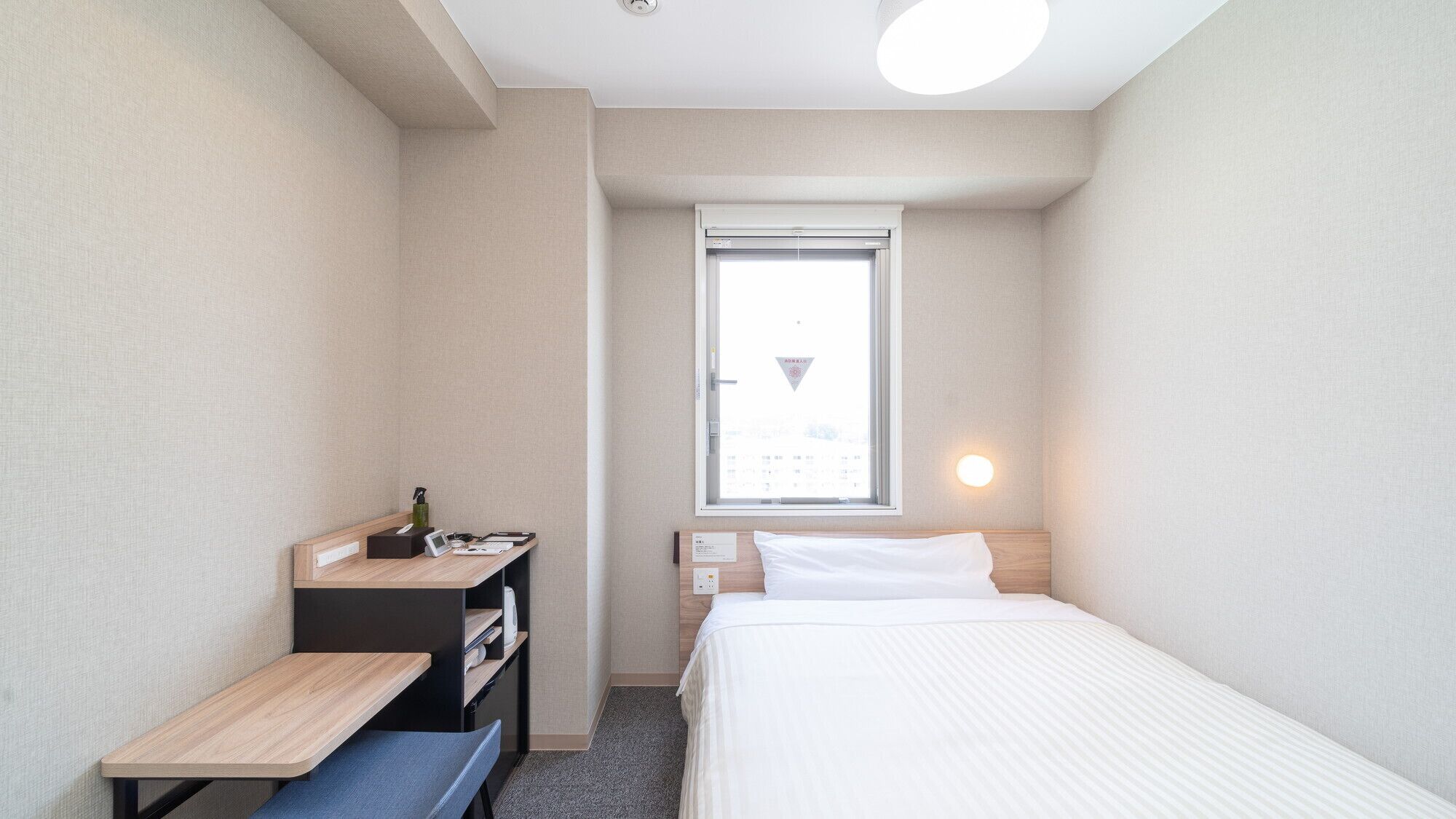 A small, well-lit hotel room with a single bed, a desk with a computer, a bench, and a window with a view.