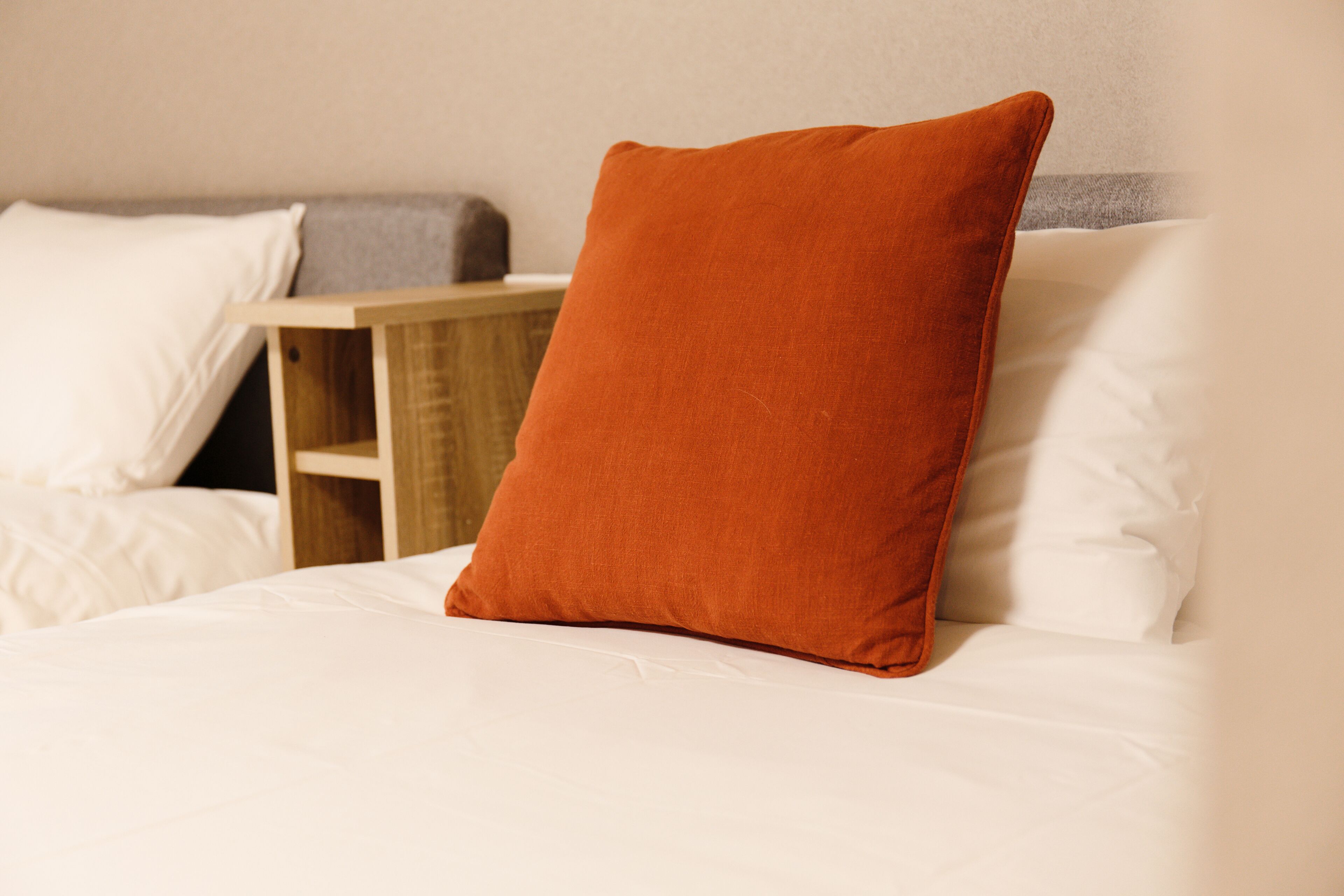 Un lit avec une literie blanche, une table de chevet en bois et un seul coussin orange.