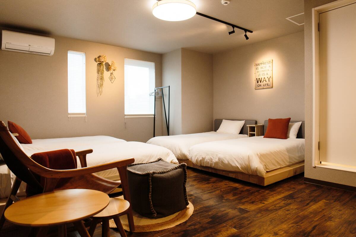 Quarto de hotel com duas camas, um espaço de estar com uma cadeira e mesa, um banquinho pequeno e um ar condicionado fixado na parede.