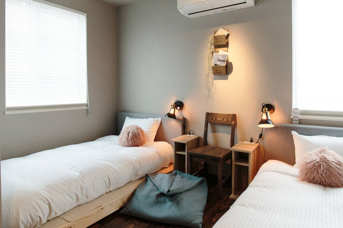 Quarto com duas camas de solteiro, uma mesa de cabeceira de madeira, uma escrivaninha e uma prateleira na parede com caixas.