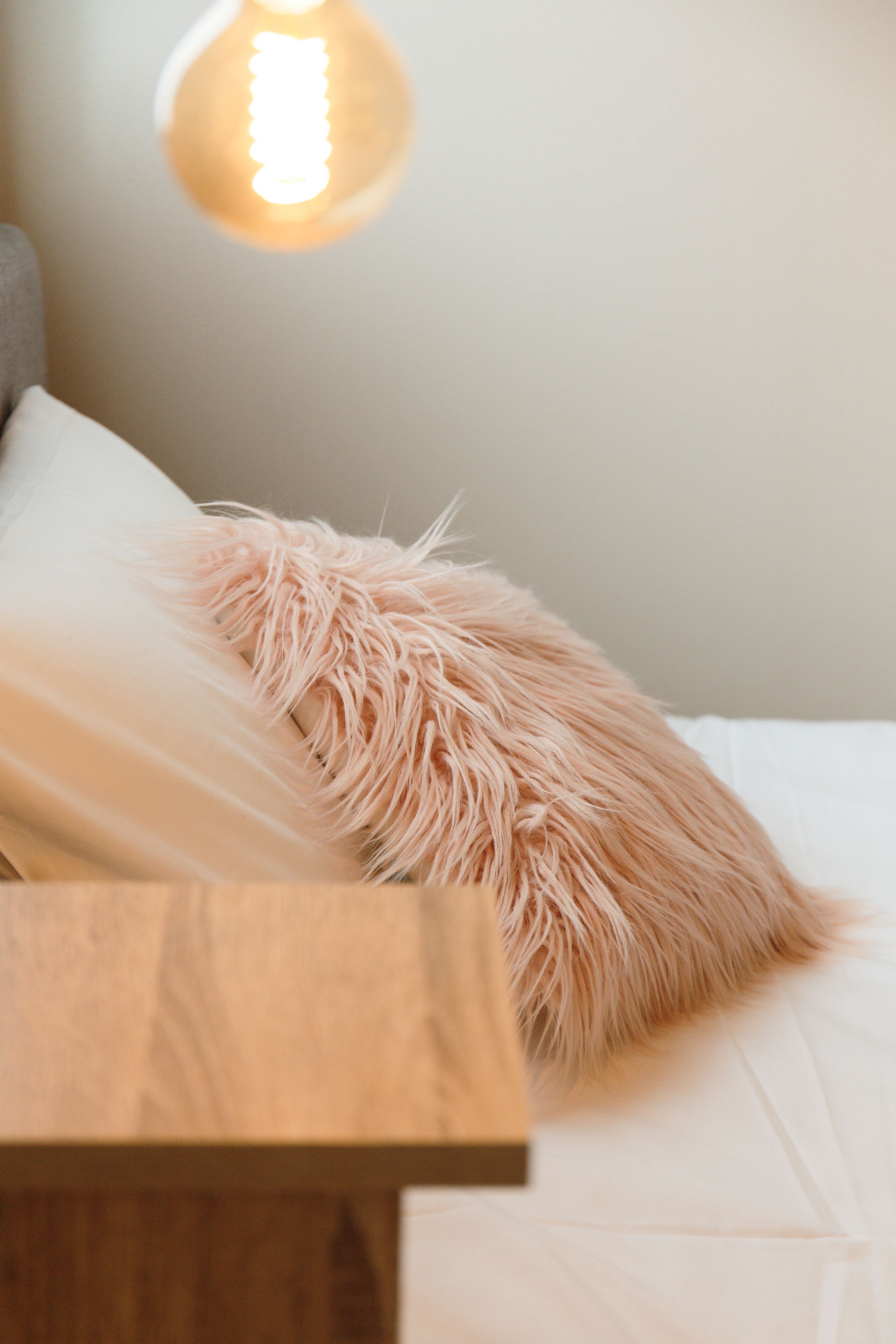 A neatly made bed with a fluffy pink pillow and a wooden bedside table.