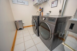 A laundry room with two front-loading washing machines, a dryer, and a small trash bin.