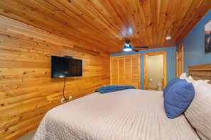 A bedroom with a wooden ceiling, a bed with a white quilt, a television mounted on the wall, and a door leading to a bathroom.