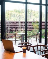 Una mesa de madera con una laptop y una taza de café, rodeada de sillas, y una vista a un espacio exterior a través de amplios ventanales.