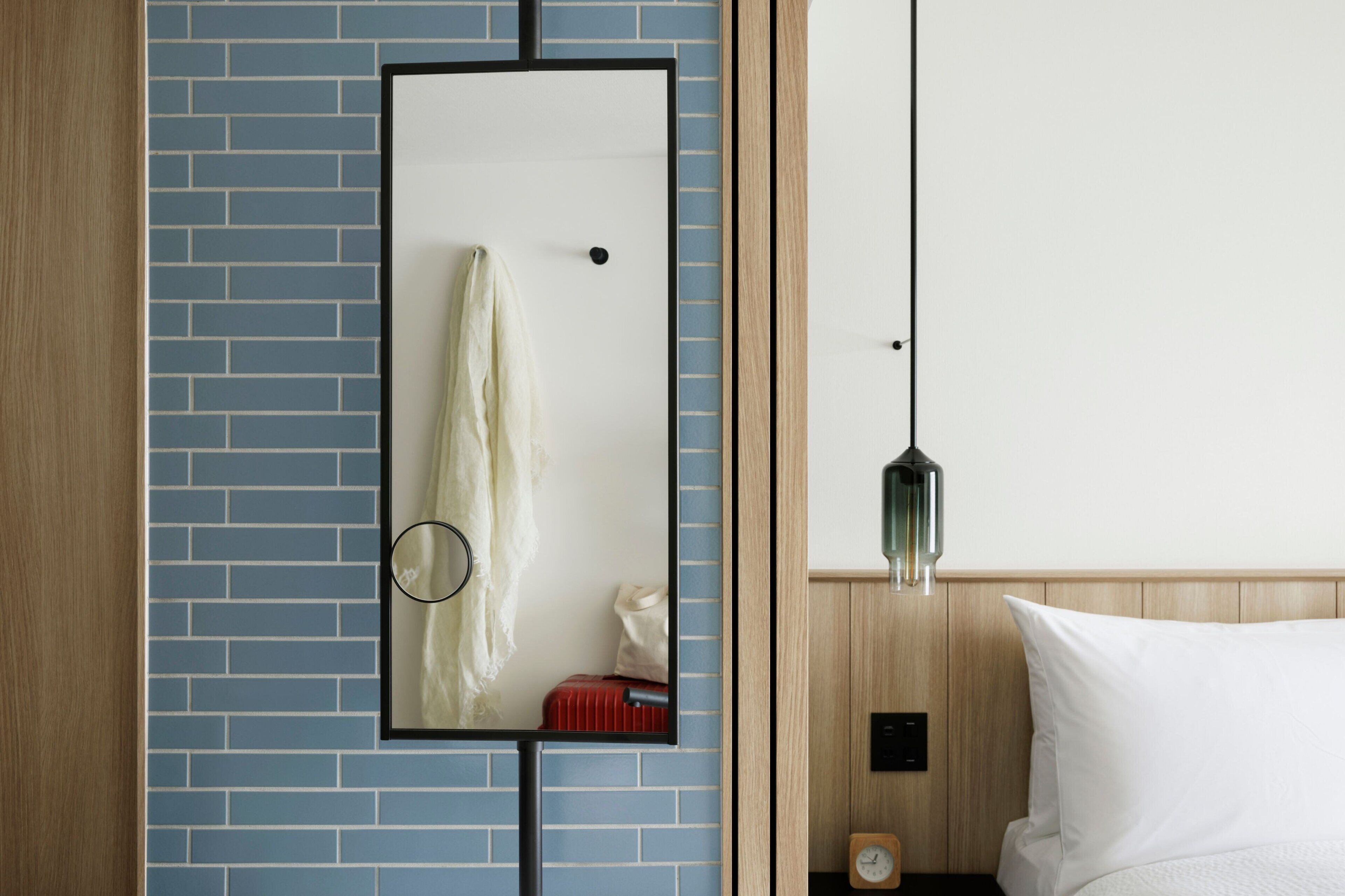 A hotel room with a blue brick wall, a mirror, a wooden headboard, a bed with white bedding, a small clock, and a hanging light fixture.