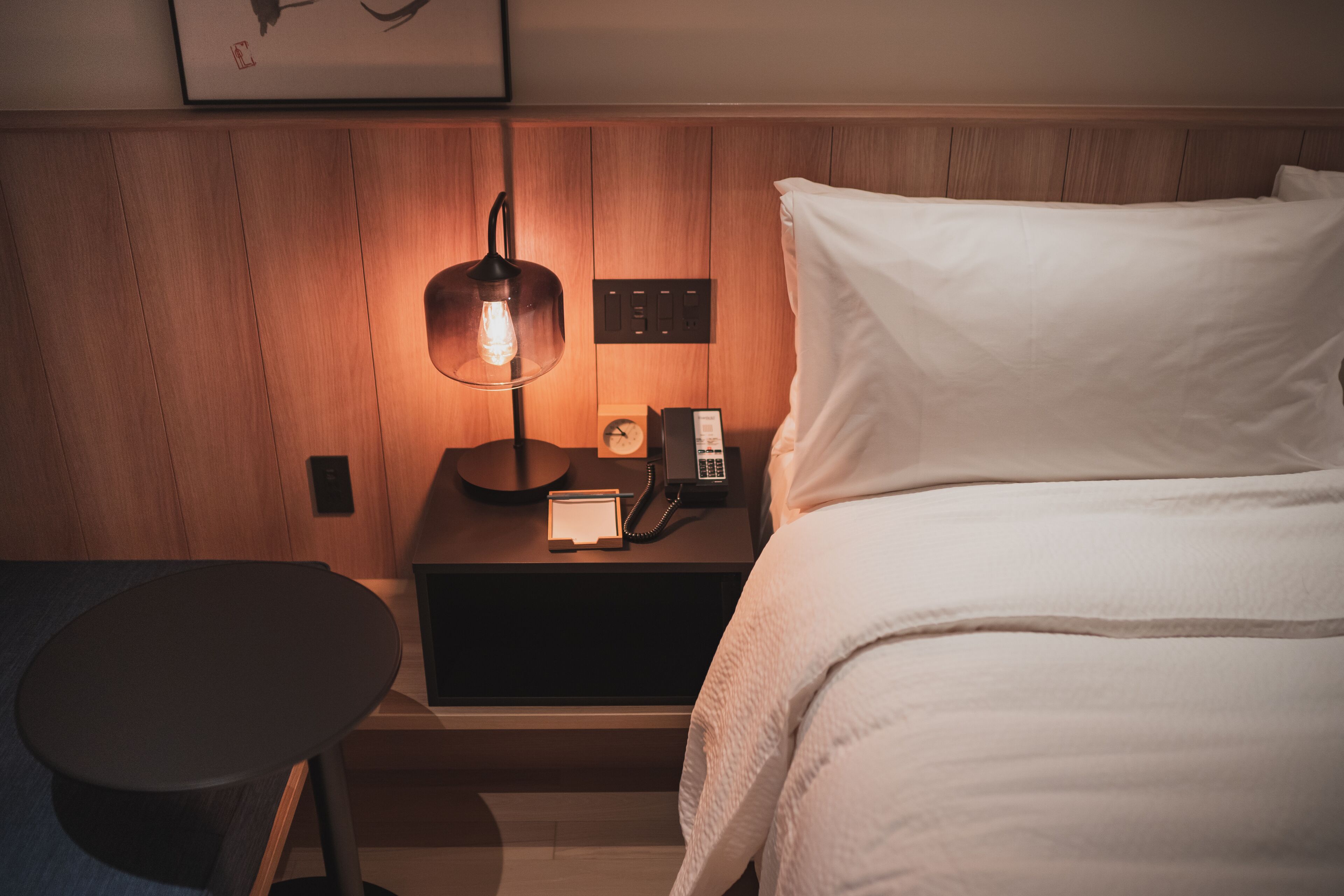 Una cama bien hecha con sábanas blancas, una mesita de noche con una lámpara, un teléfono y un pequeño reloj sobre un mueble de madera.