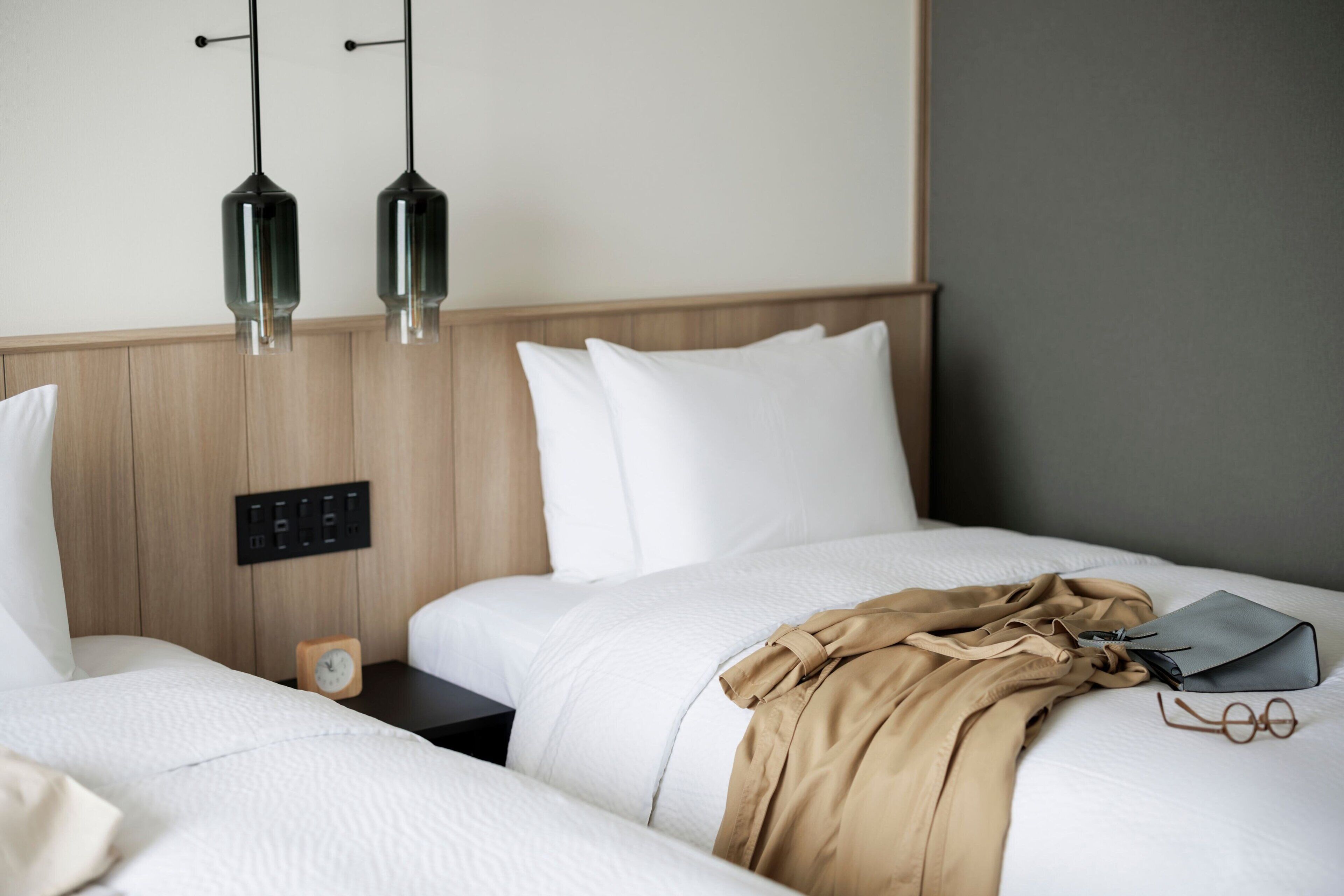 A hotel room with two beds, a wooden headboard, a bedside table with a clock, and two pendant lights.