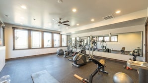 Een fitnessruimte met diverse trainingsapparaten, grote ramen en een plafondventilator.