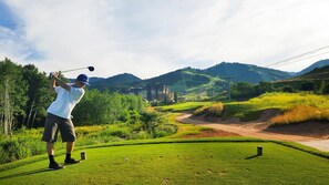 Un campo da golf con una persona con camicia bianca e pantaloncini scuri che si prepara a colpire una pallina da golf.