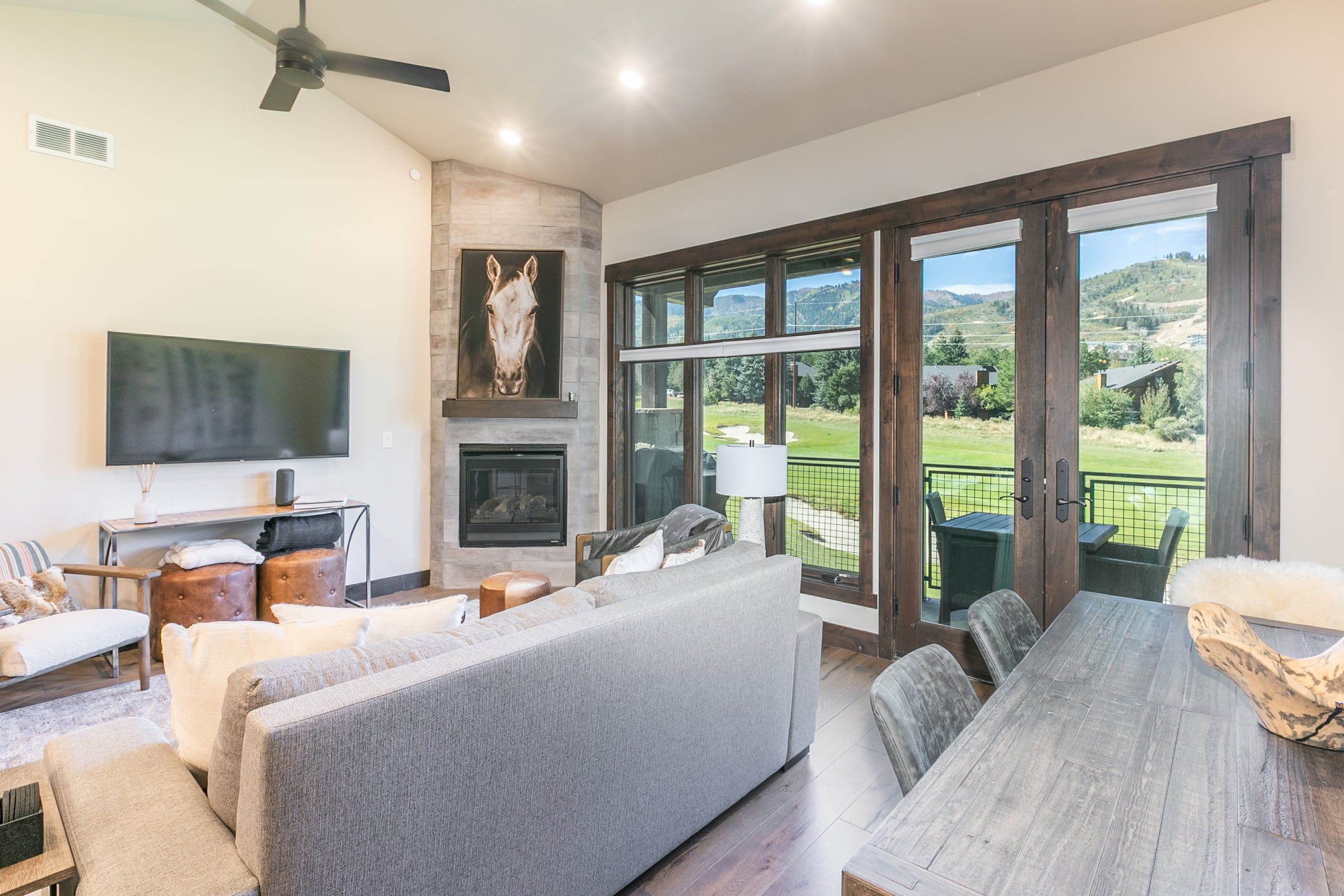 A spacious living room with a large gray sofa, a flat-screen TV mounted on the wall, a fireplace, and a view of a green landscape through large windows.