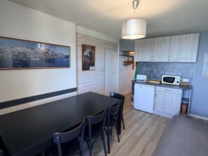 A modern kitchen with white cabinets, a black dining table, and chairs. There is a painting on the wall and a ceiling light fixture.