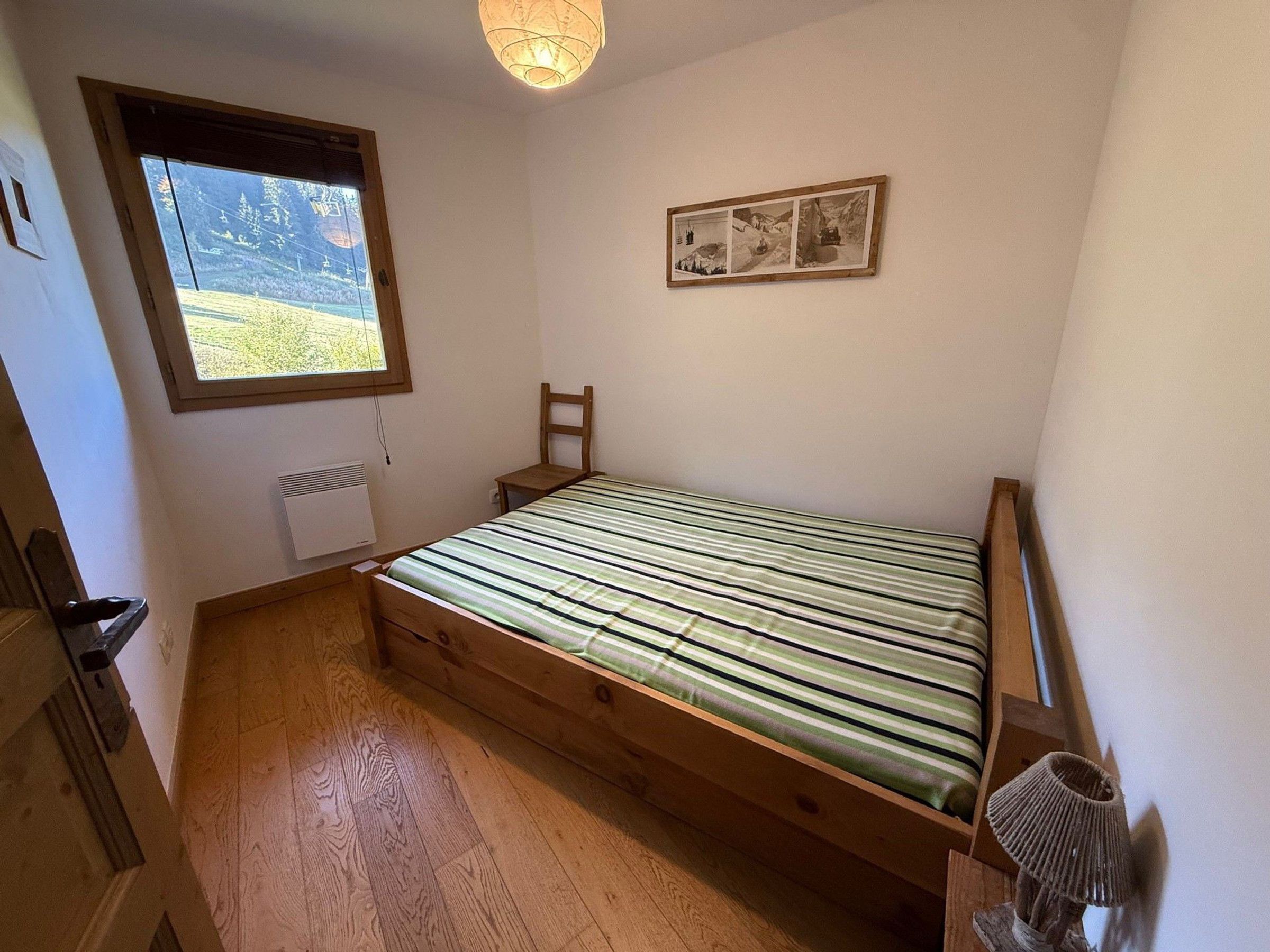 A small, single-bed room with a wooden floor, a window with a view of greenery, a radiator, and a framed picture on the wall.