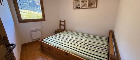 A small, single-bed room with a wooden floor, a window with a view of greenery, a radiator, and a framed picture on the wall.