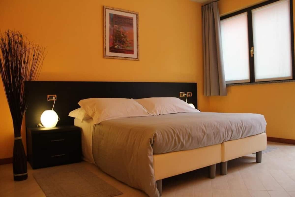 A bedroom with a bed, nightstands, a lamp, a picture on the wall, and a window with curtains.