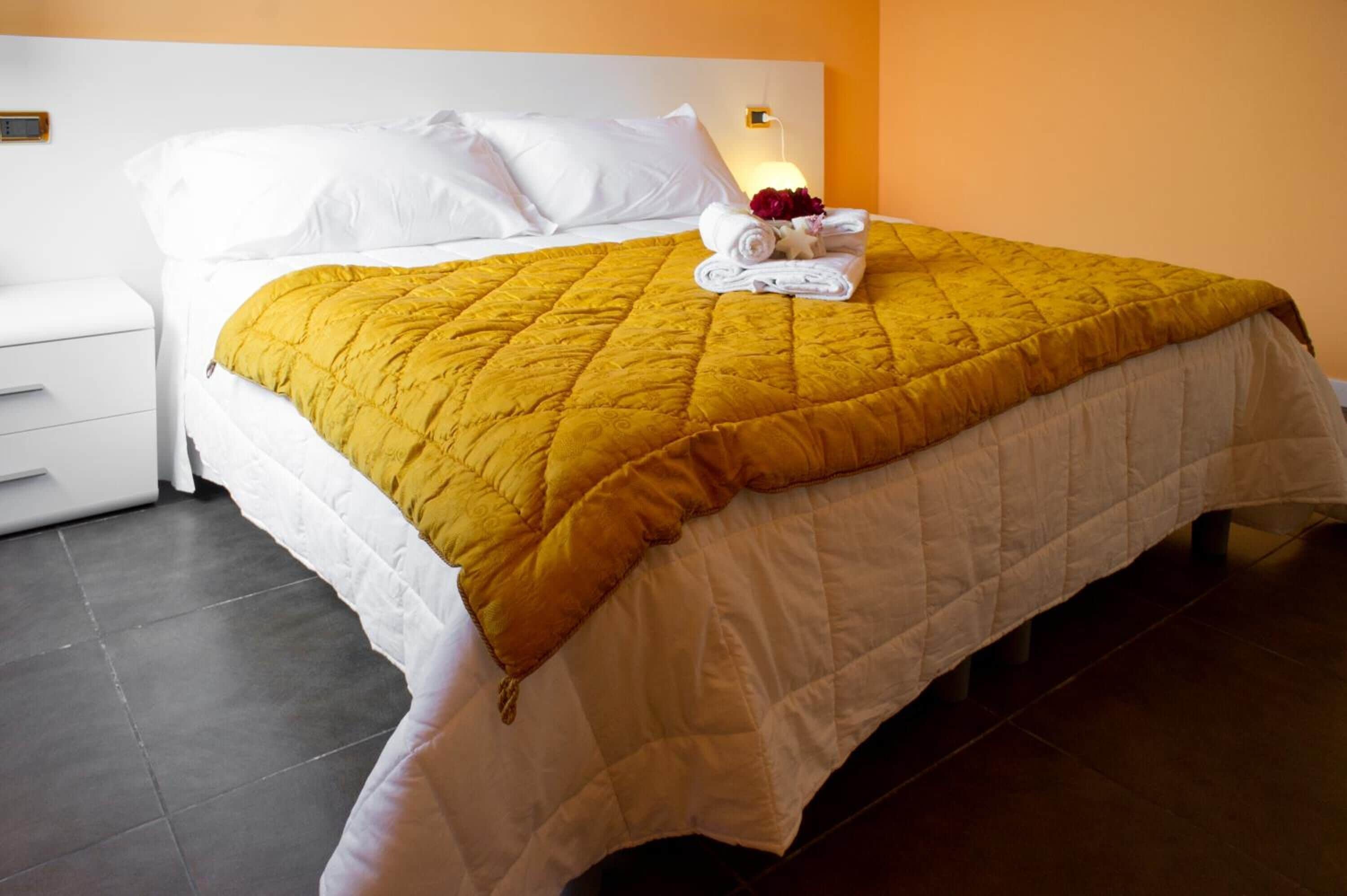 A neatly made bed with a white headboard and a yellow quilt, a white nightstand with a drawer, and a wall-mounted light fixture.