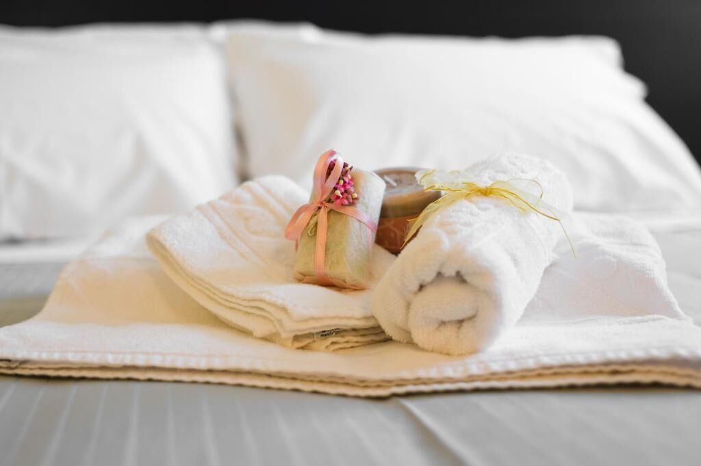 Una toalla blanca doblada con un lazo rosa y una pequeña botella sobre una cama con ropa blanca.