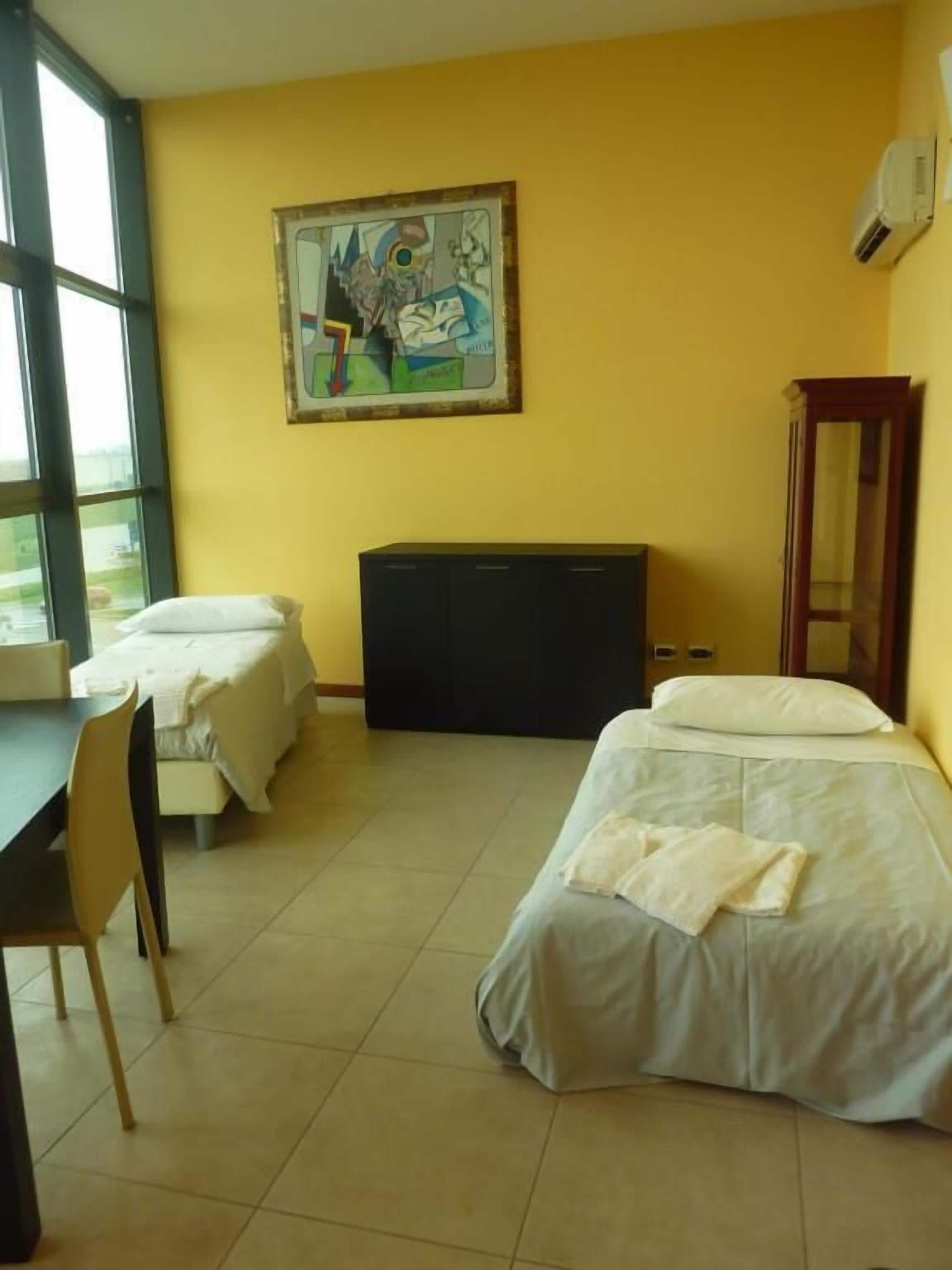 Una camera d'albergo con due letti, una scrivania, una sedia, un'ampia finestra e un quadro appeso al muro.