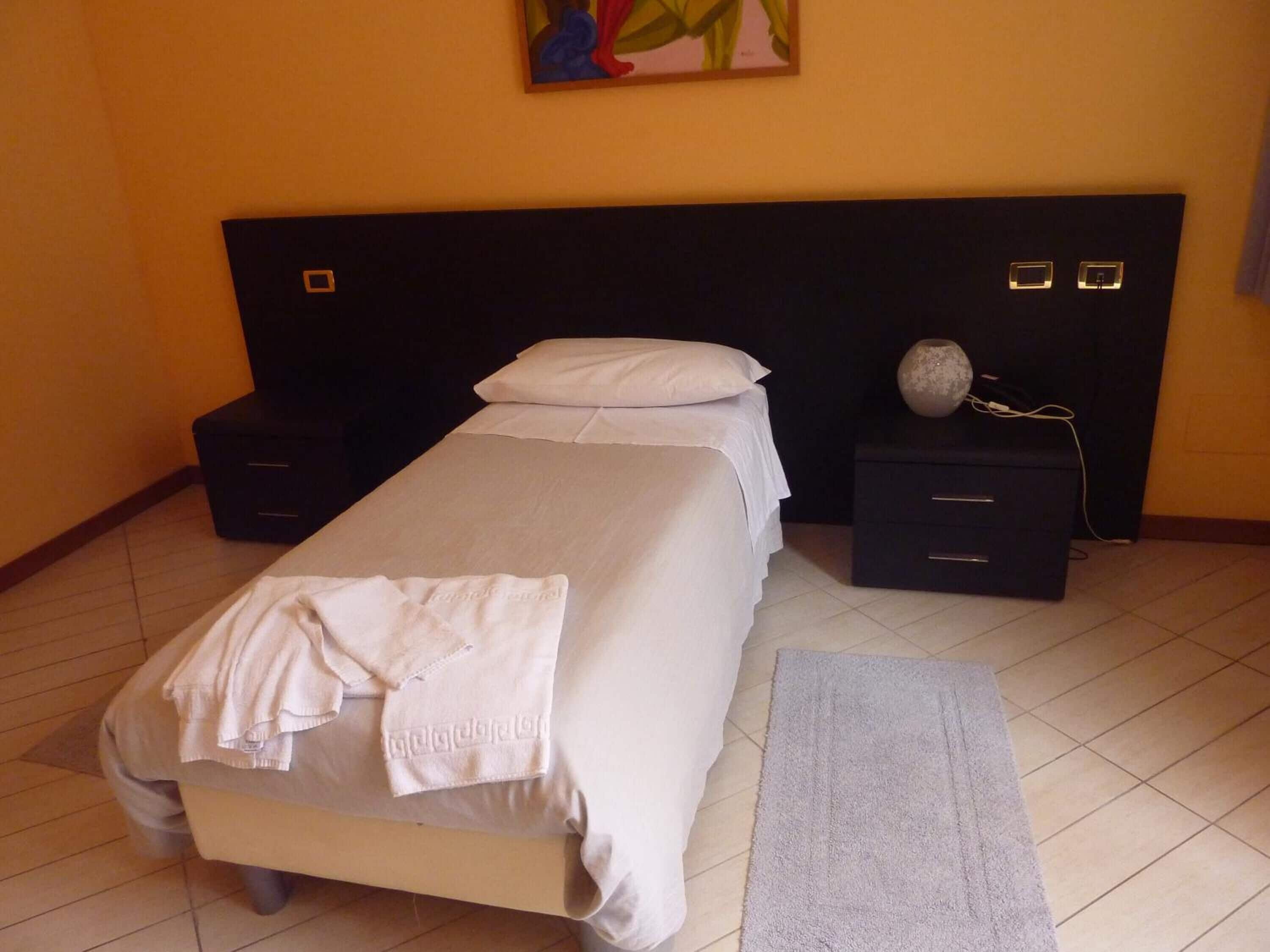 Una cama bien hecha con una colcha blanca y una toalla doblada, una mesita de noche con un florero, y un cabecero con dos interruptores de luz.