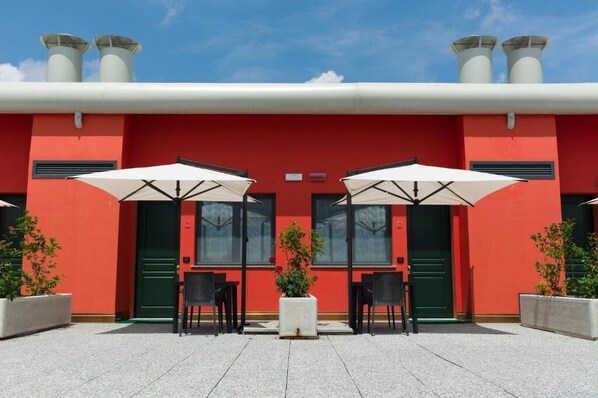 A red building with two green doors, two white umbrellas, and two black chairs.