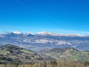 Vista panorâmica de uma paisagem montanhosa com picos nevados, céu azul límpido e um vale com edifícios dispersos.