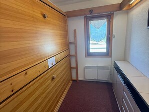 Quarto pequeno com guarda-roupa de madeira, radiador, janela com vista e armário branco.