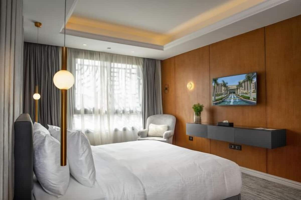 A hotel room with a bed, a chair, a TV mounted on a wooden panel, and a window with curtains.
