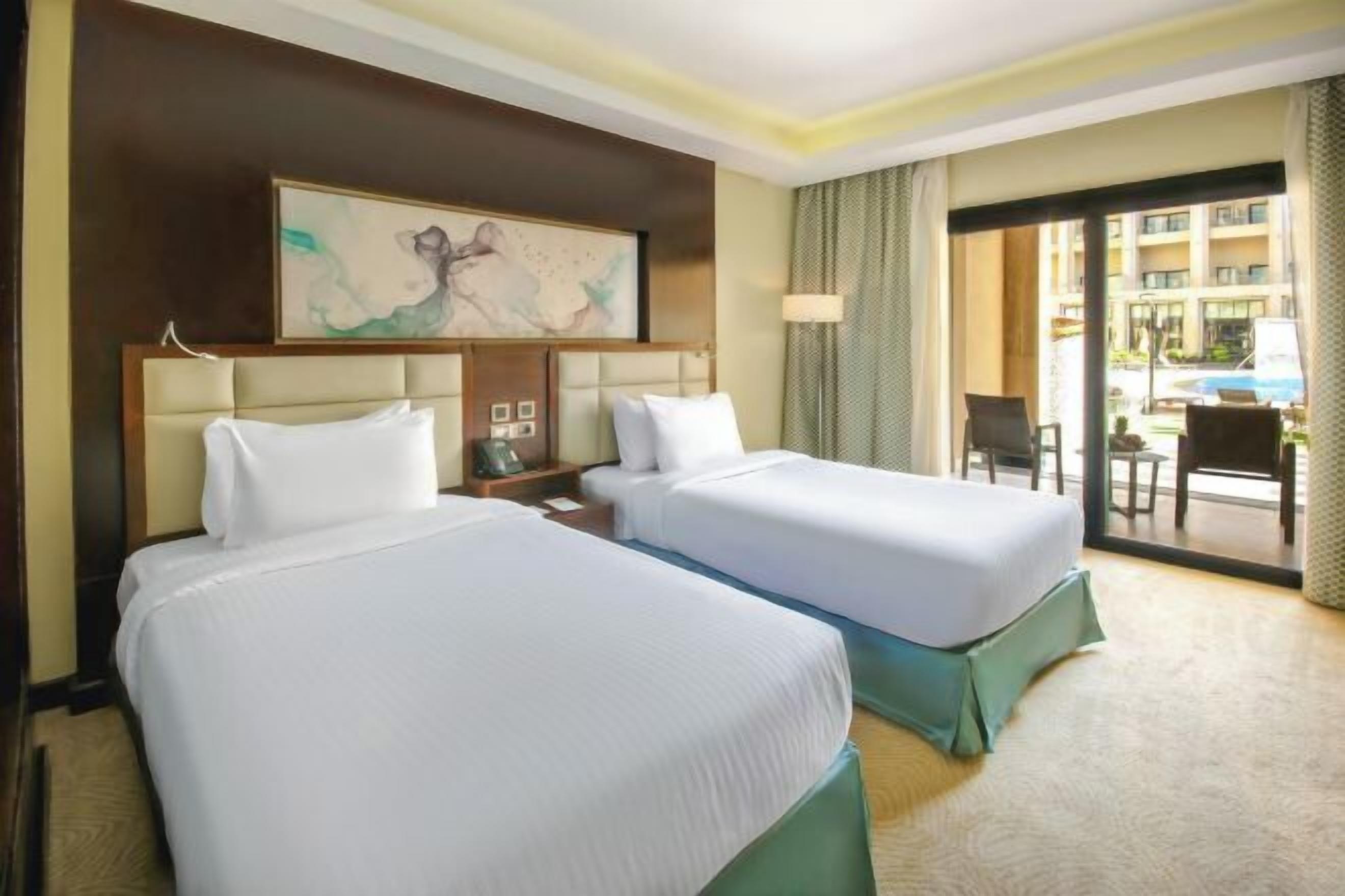 Habitación de hotel con dos camas, un ventanal grande y vistas a la piscina y edificios exteriores.