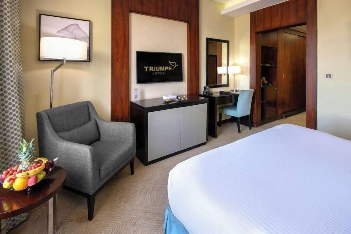 A hotel room with a bed, a desk, a grey armchair, a fruit bowl, a television, and a wardrobe.