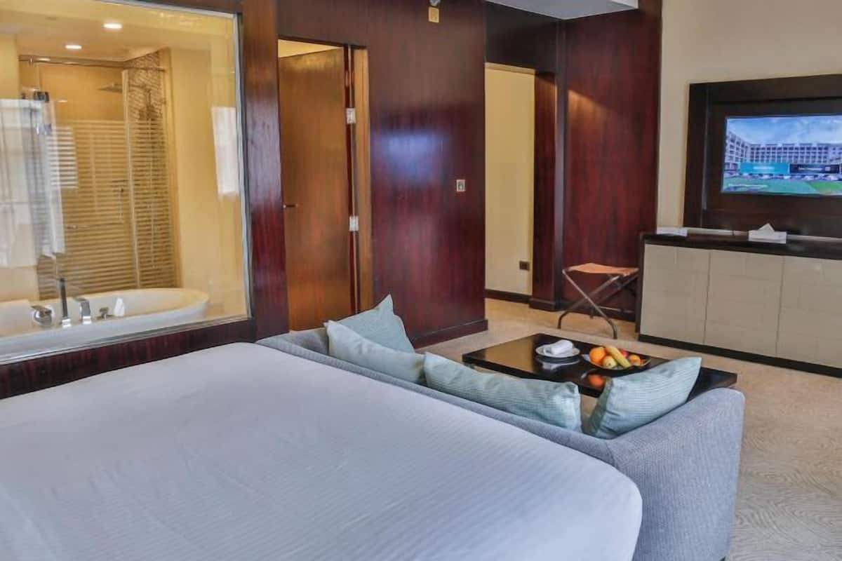 A hotel room with a large bed, a sofa, a small table with fruit, a TV, and a bathroom visible through a glass door.