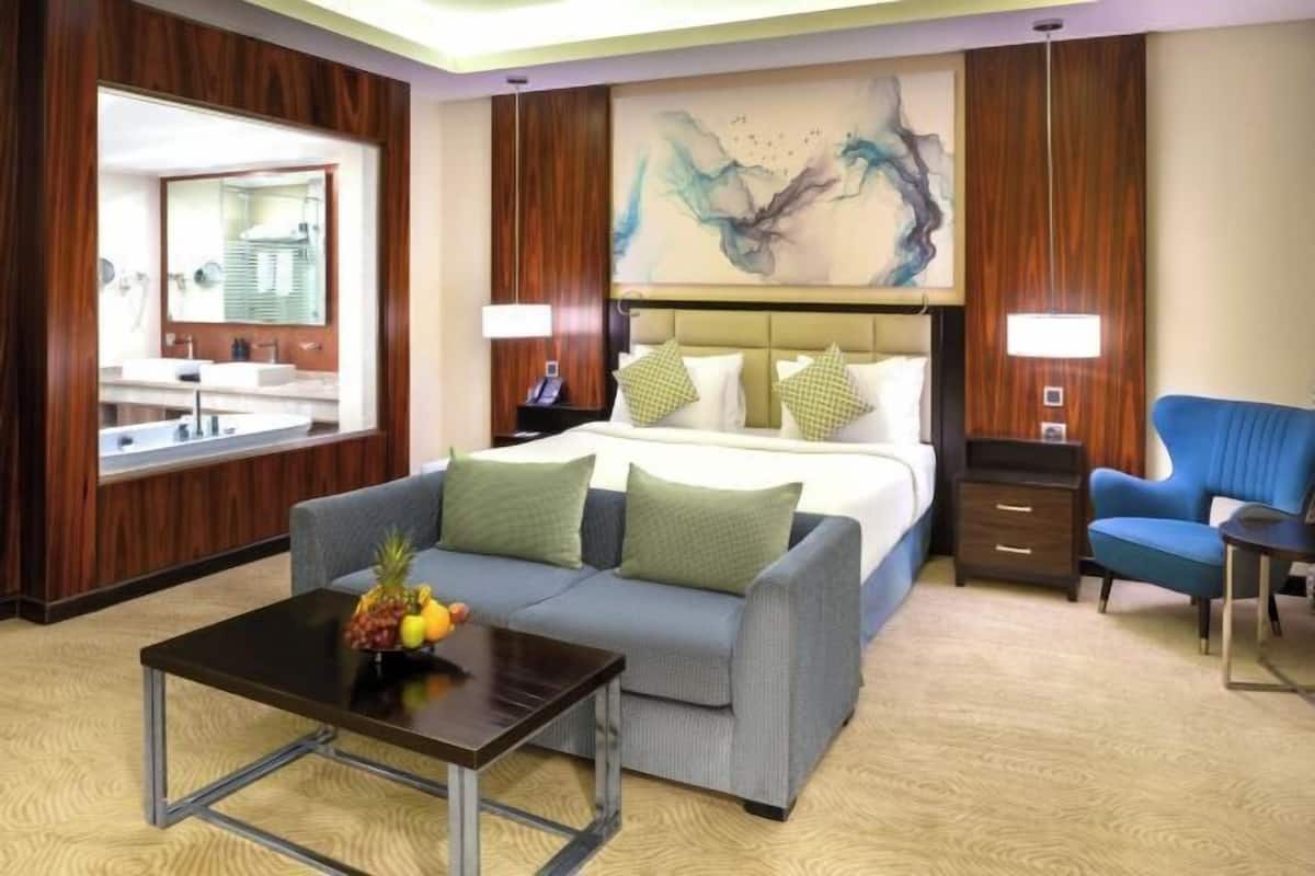 A hotel room with a bed, a sofa, a coffee table with fruit, a desk, and a mirror.