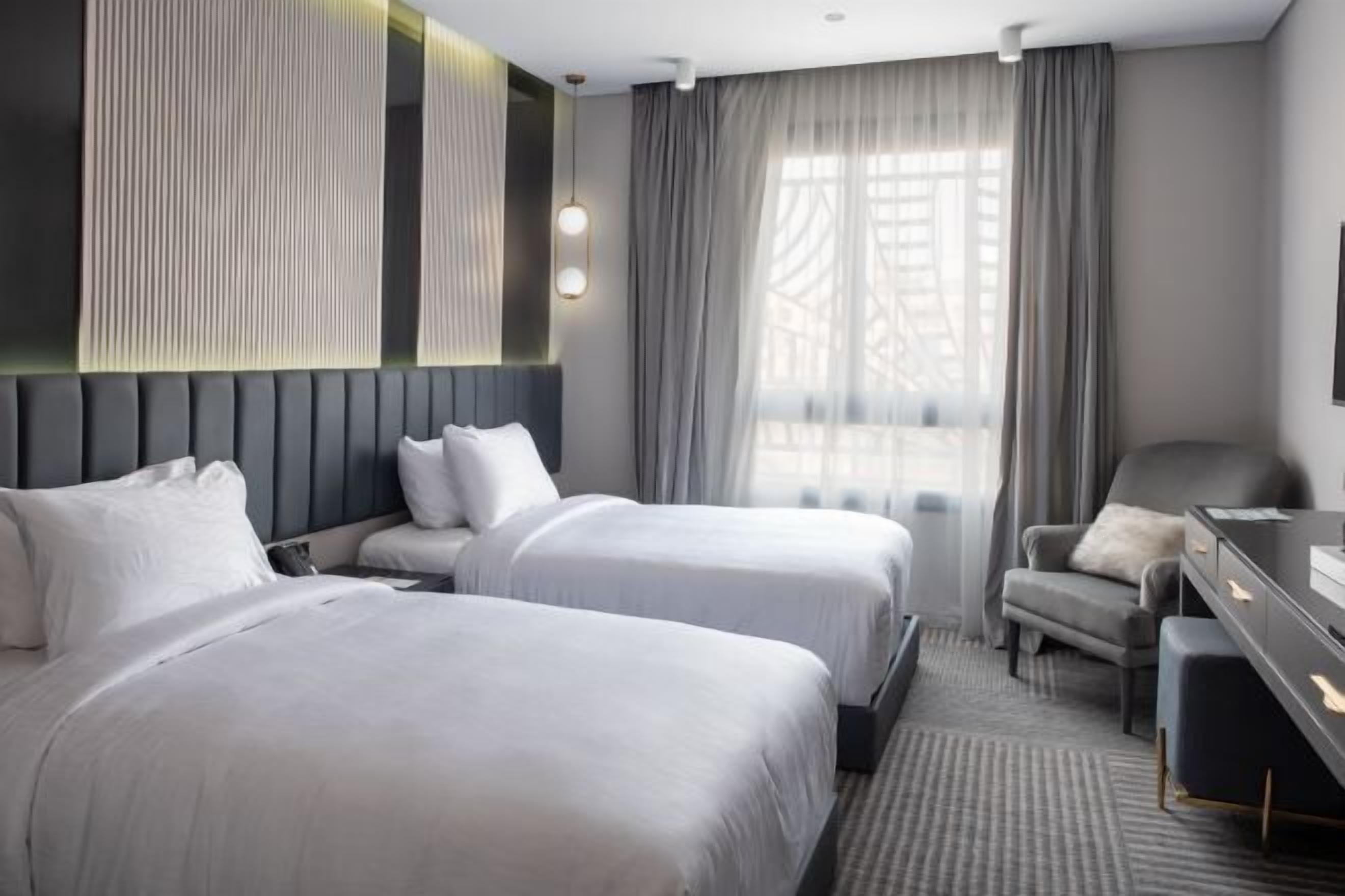 Habitación de hotel con dos camas, un escritorio, una silla y un ventanal con cortinas.