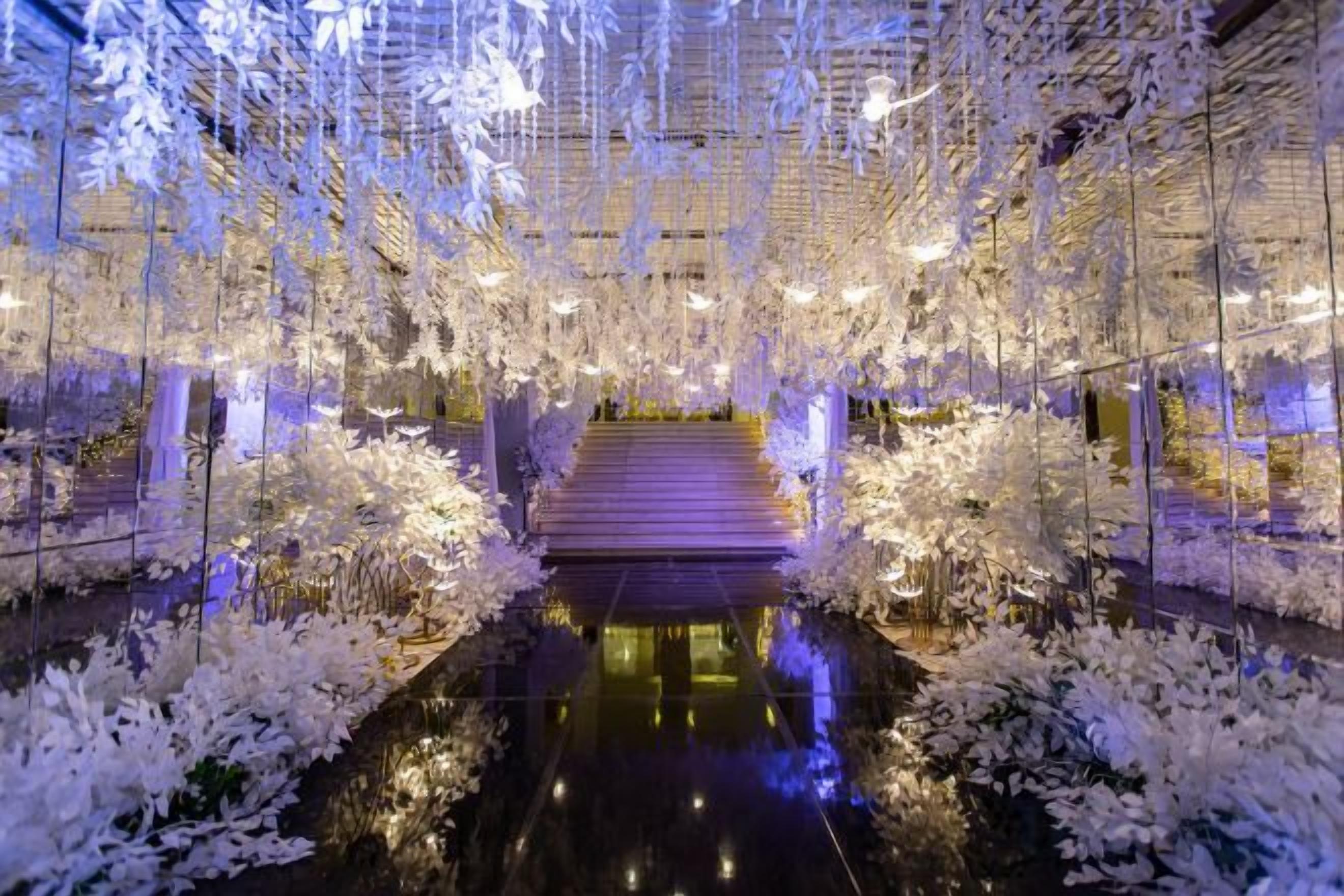 Un espacio interior con piso reflectante, una escalera central y decoraciones florales colgantes.