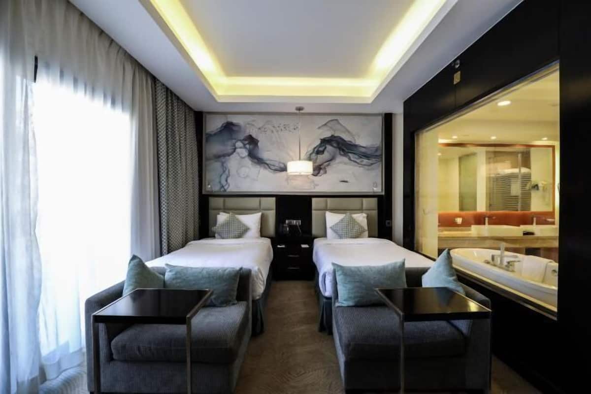 A hotel room with two beds, a large artwork above the beds, a sitting area with a table and chairs, and a large window with curtains.