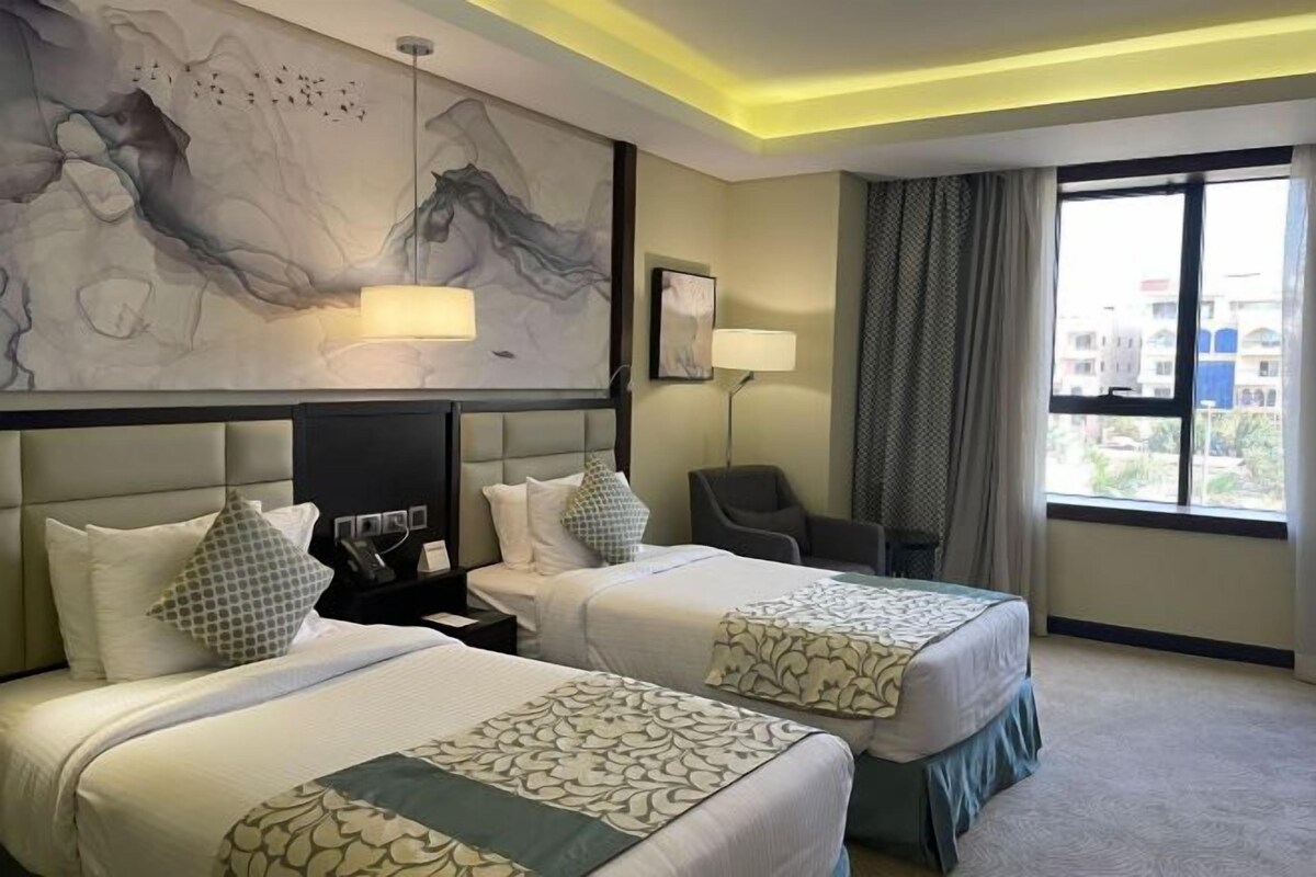 Ett hotellrum med två sängar, ett skrivbord, en stol, ett stort fönster och abstrakt konst på väggen.