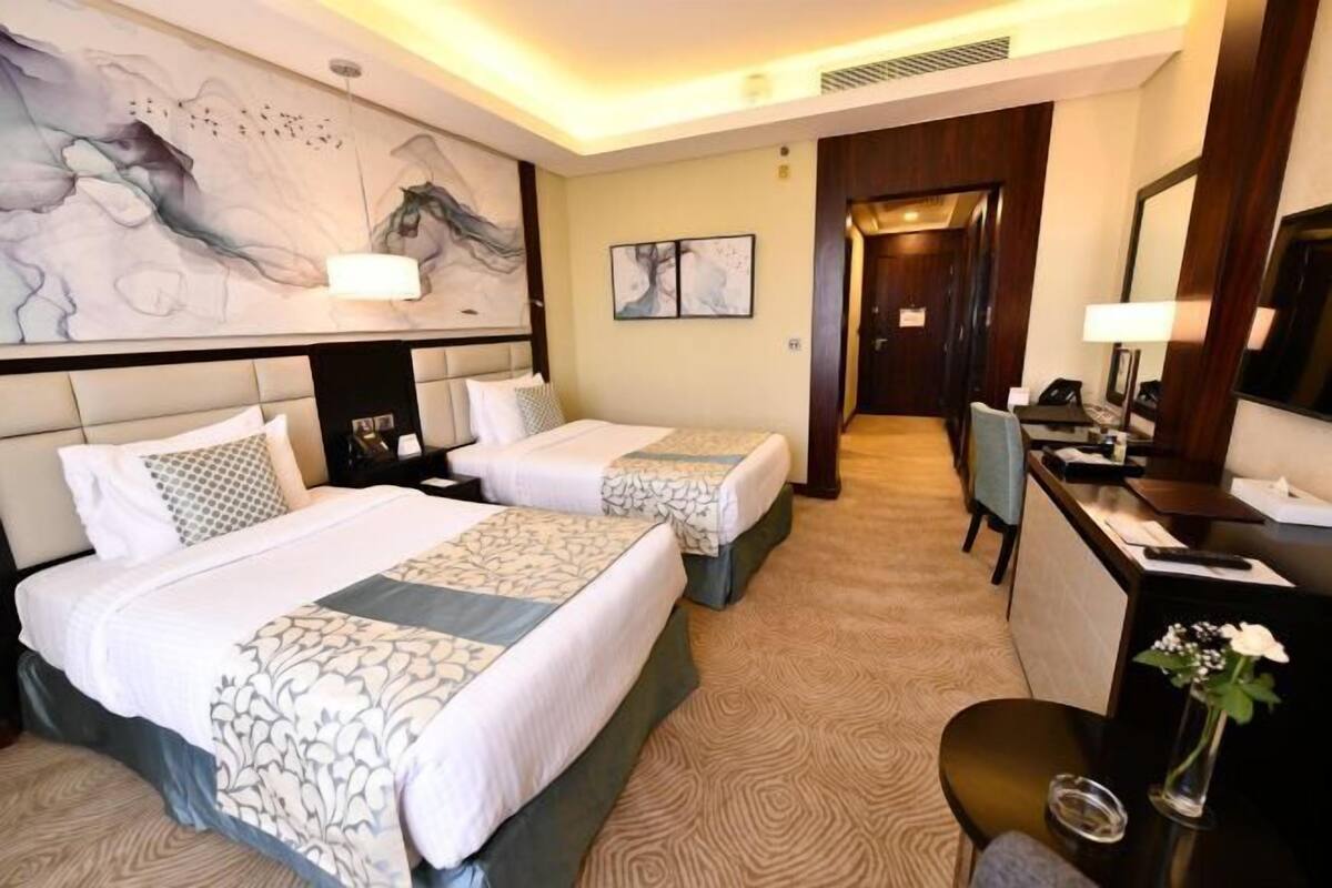 A hotel room with two beds, a desk, a chair, a desk lamp, a mirror, and artwork on the walls.