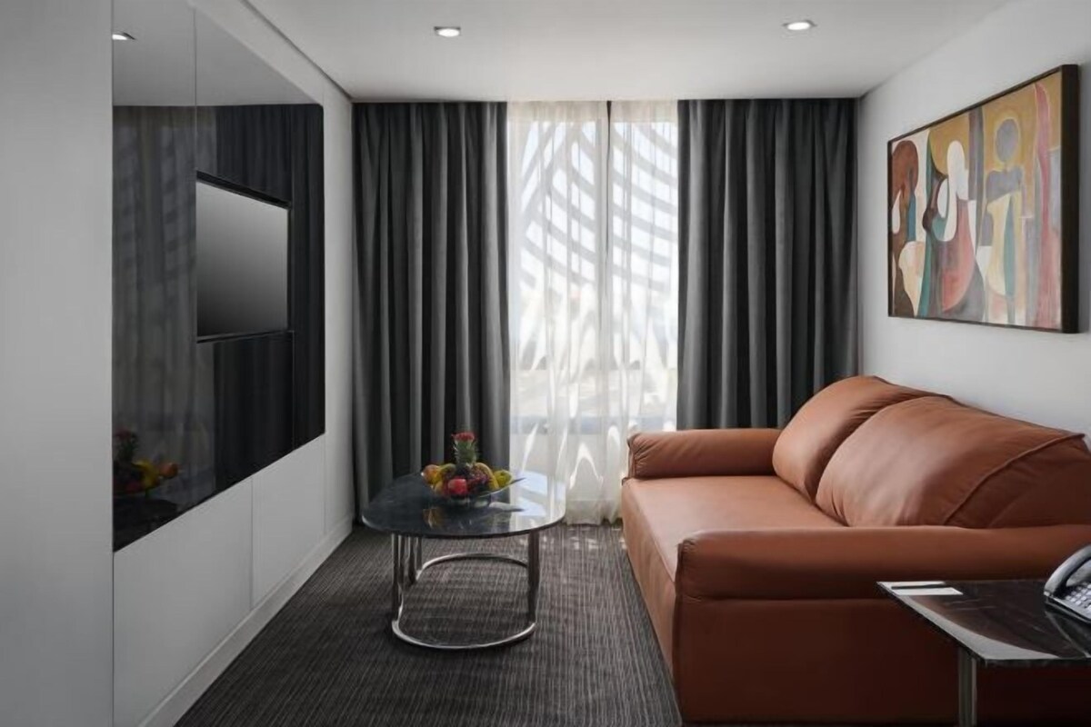 A modern hotel room with a sofa, a small round table with fruit, a flat-screen TV, and a painting on the wall.