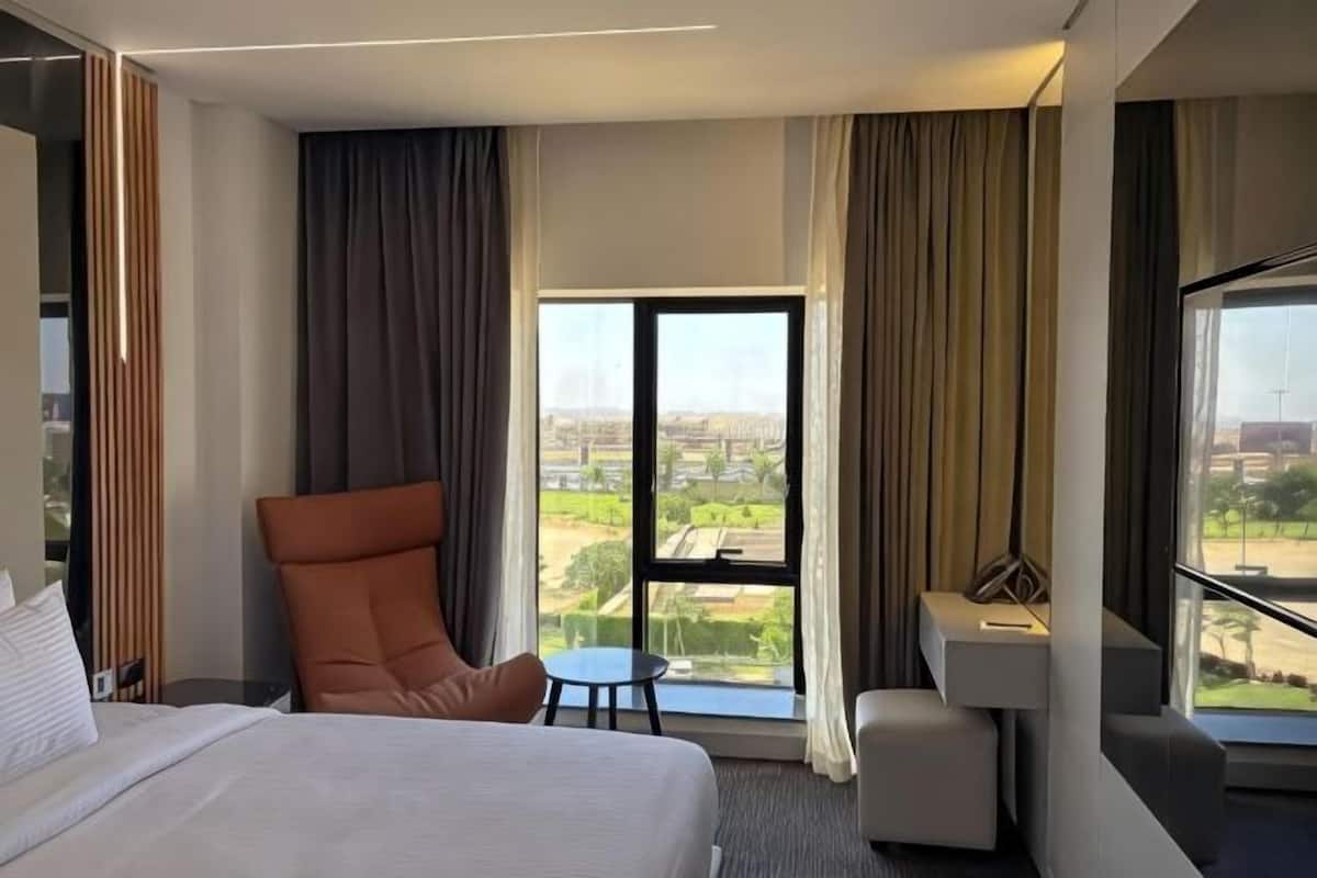 A hotel room with a bed, a chair, a small table, and a view of the city through large windows.