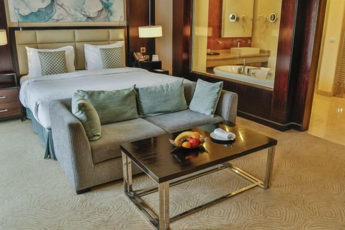 A hotel room with a bed, a sofa, a coffee table with fruit, a bathroom with a mirror, and a decorative wall.