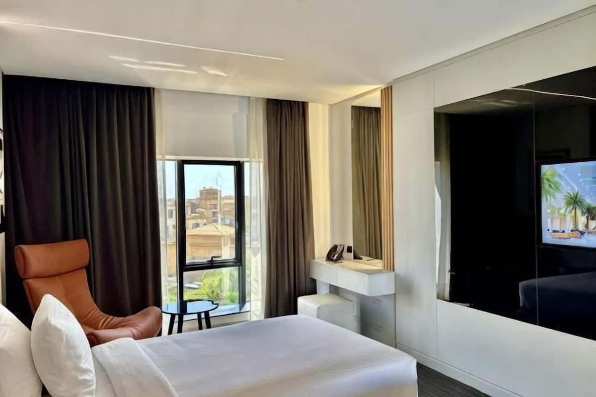 A hotel room with a bed, a chair, a small table, a desk, and a large window with a view.
