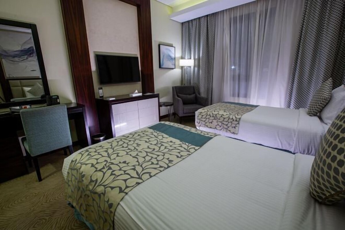 A hotel room with two beds, a desk, a chair, a television, and a window with curtains.