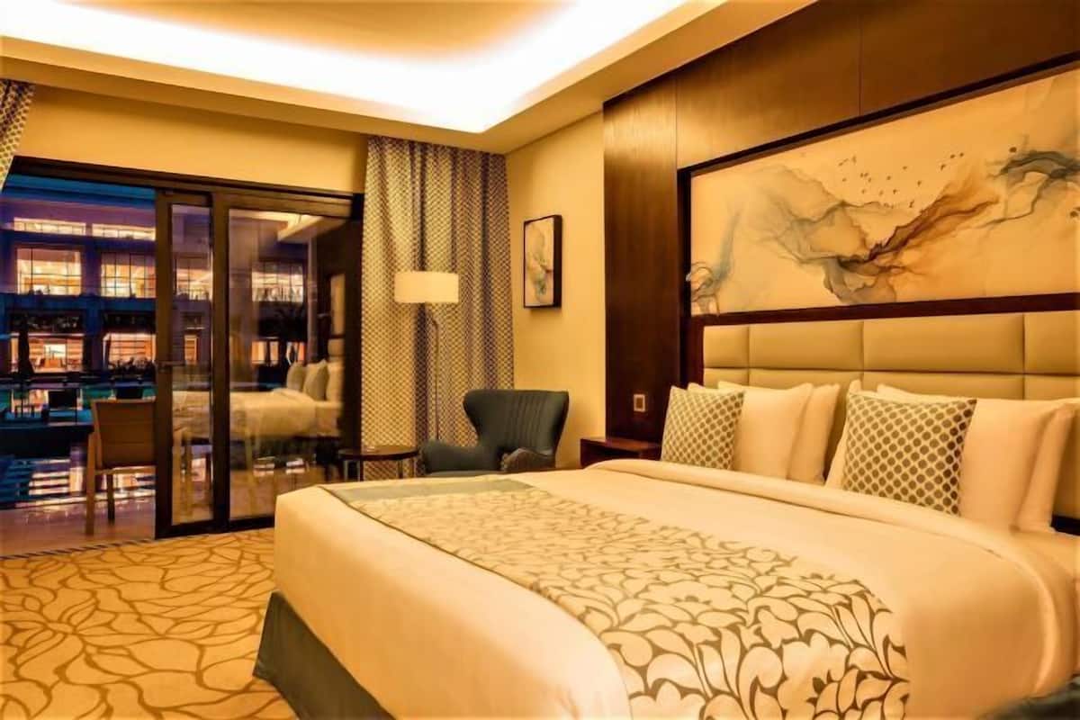 A hotel room with a large bed, a chair, a desk, and a view of the hotel's interior.