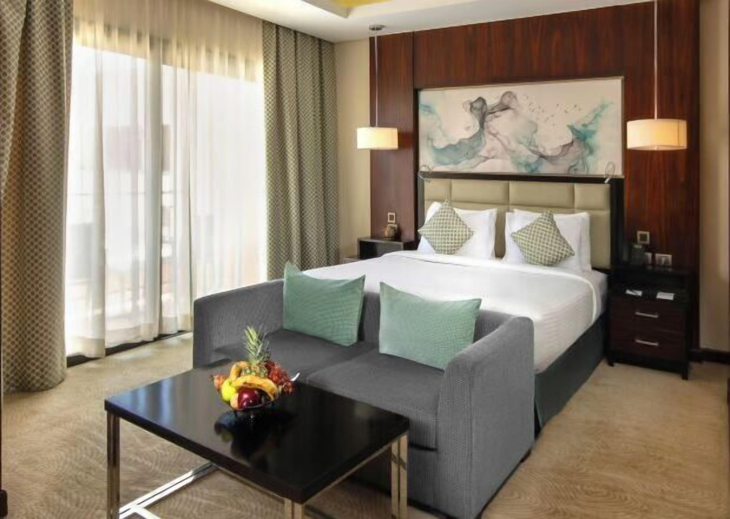 Habitación de hotel con cama, sofá, mesa de centro con fruta y un cuadro en la pared.
