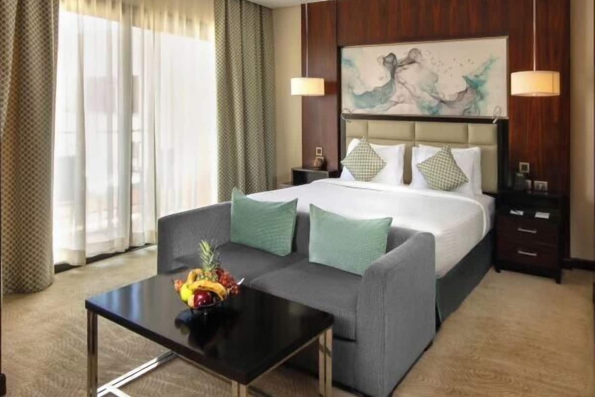 A hotel room with a bed, a sofa, a coffee table with fruit, and a painting on the wall.