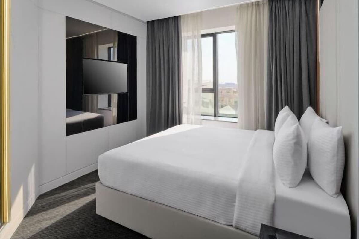 A modern hotel room with a large bed, a flat-screen TV mounted on the wall, and a view of the city through the window.