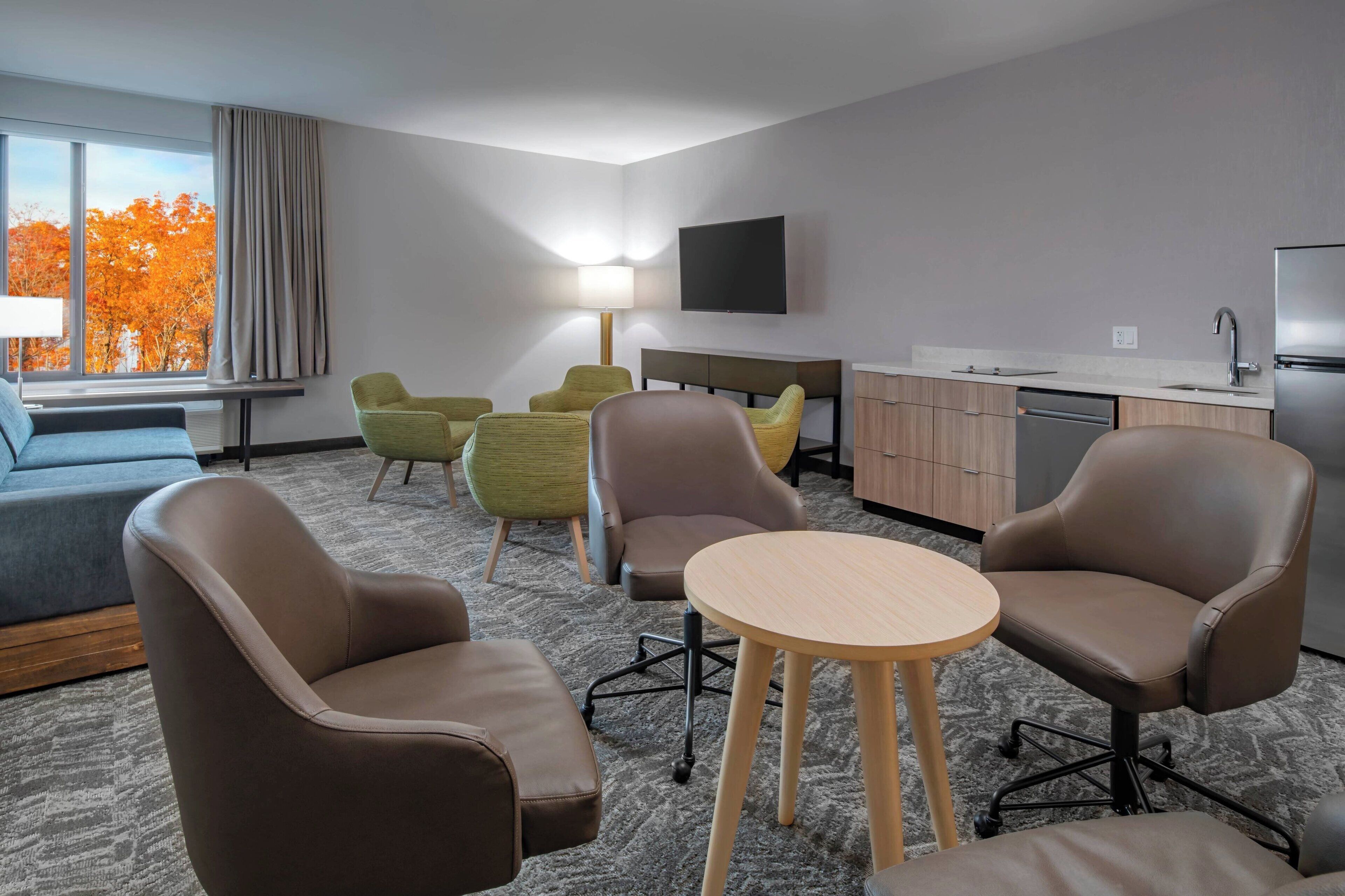 Una habitación de hotel moderna con un sofá, dos sillones, una mesita, televisor, escritorio con lámpara y zona de cocina.