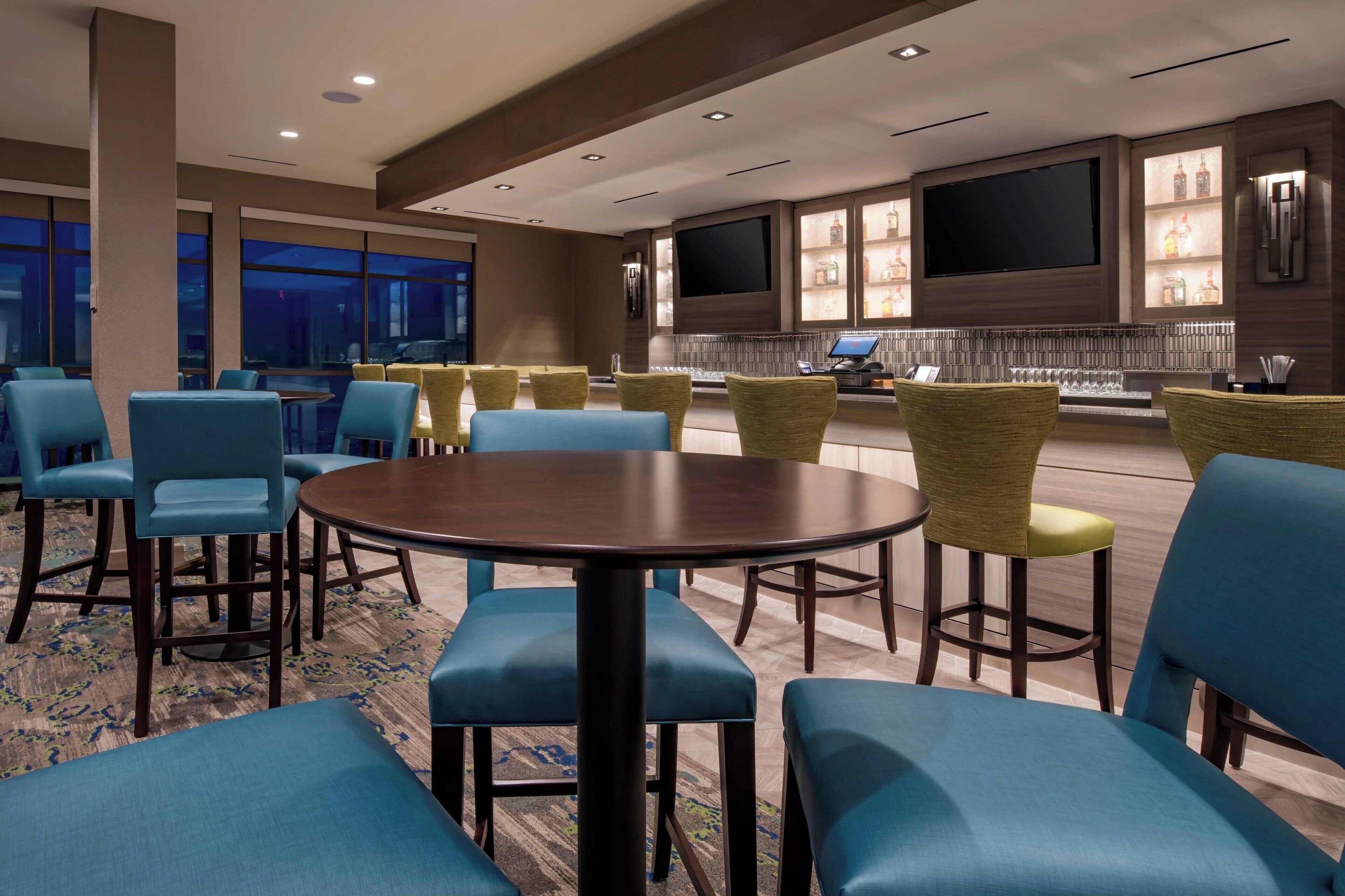 A hotel lobby with a large round table, blue and yellow chairs, a TV, and a bar area with bottles.